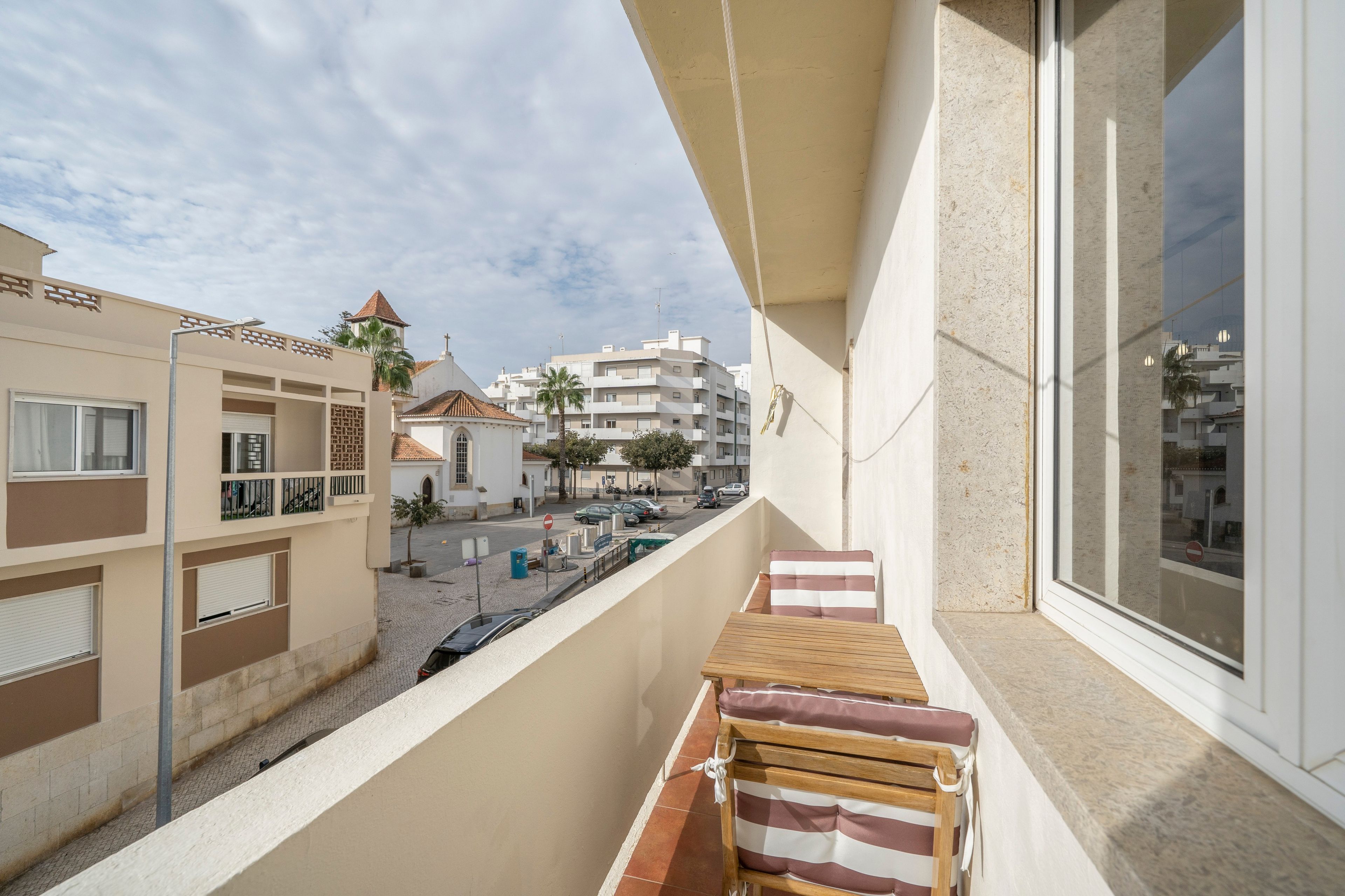 Ein Balkon mit einem Holzstuhl und Blick auf eine Straße mit Gebäuden und Palmen.