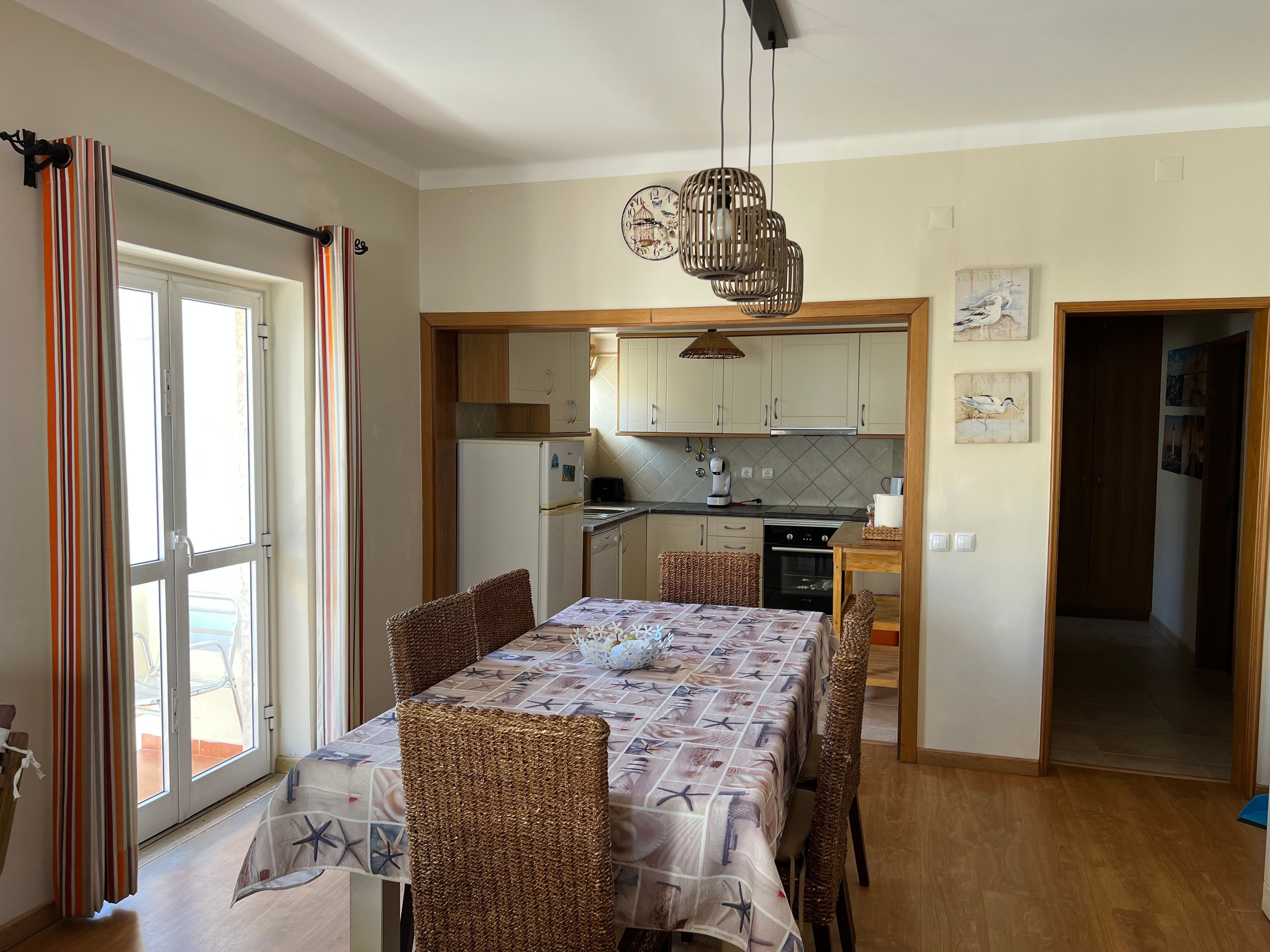 Cozinha compacta com eletrodomésticos modernos, área de refeições com cadeiras de vime e vista através de uma porta de vidro.