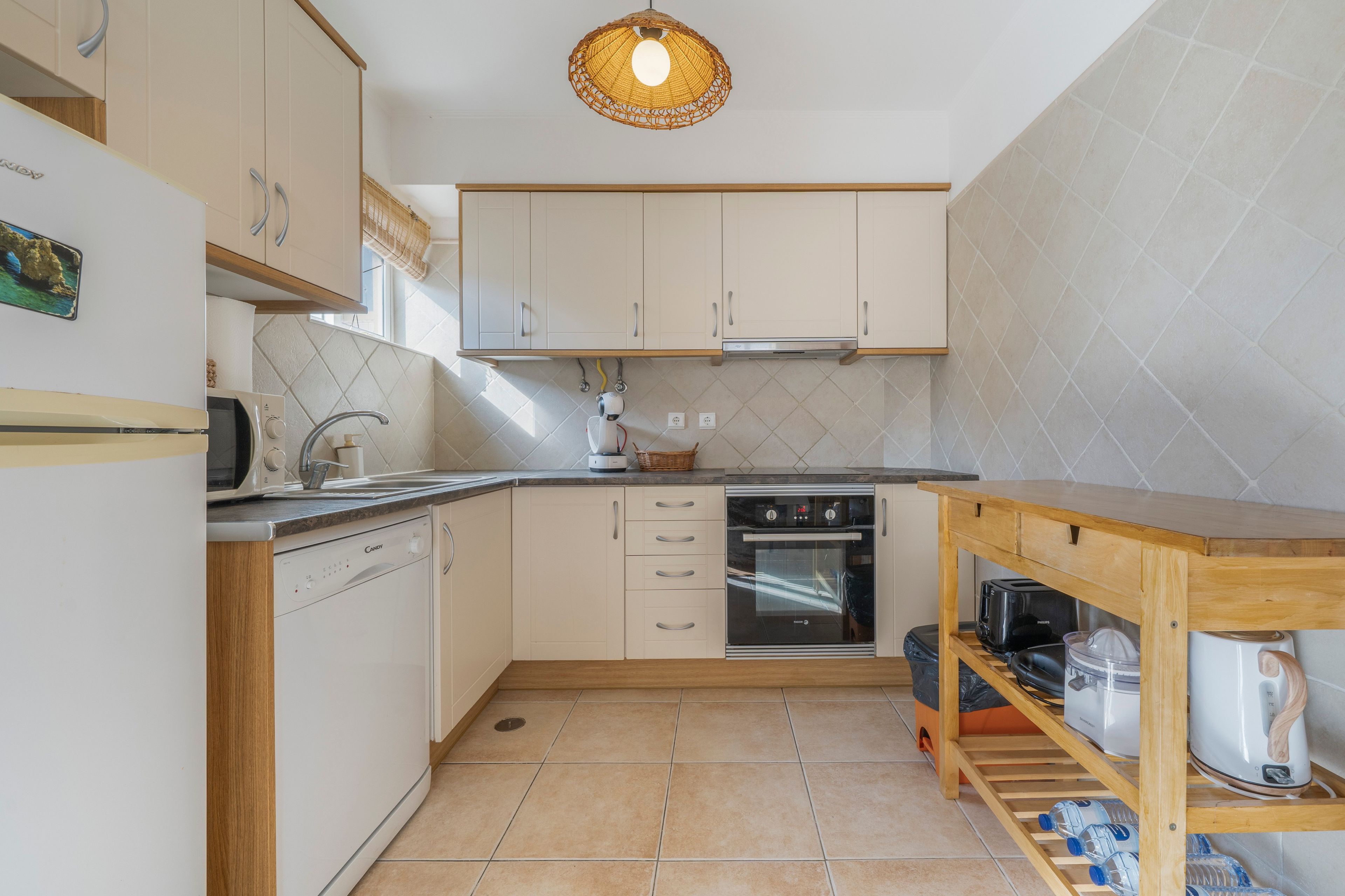 Una cocina compacta con armarios blancos, lavavajillas, horno y una mesa de madera con hervidor.