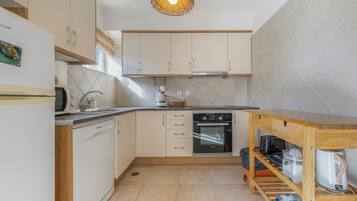 Una cocina compacta con armarios blancos, lavavajillas, horno y una mesa de madera con hervidor.