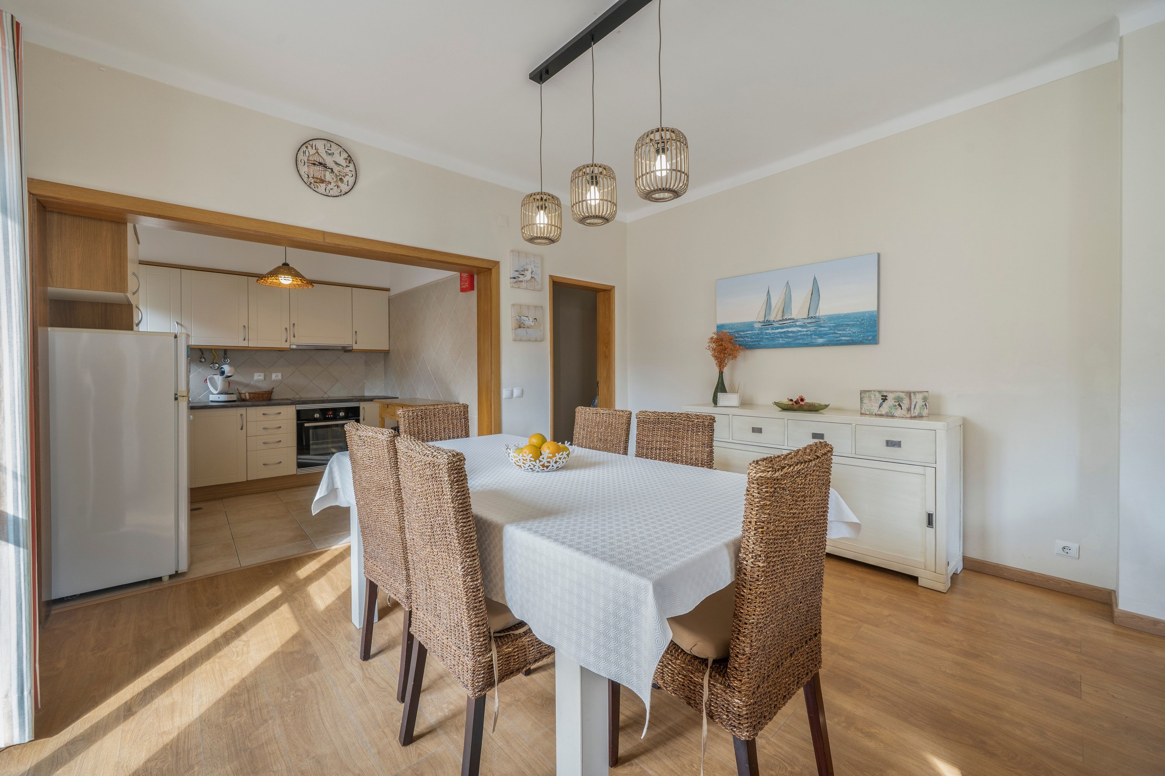 Un'area pranzo con un tavolo apparecchiato per sei persone, circondato da sedie in vimini, in una stanza con pavimento in legno e vista sulla cucina sullo sfondo.