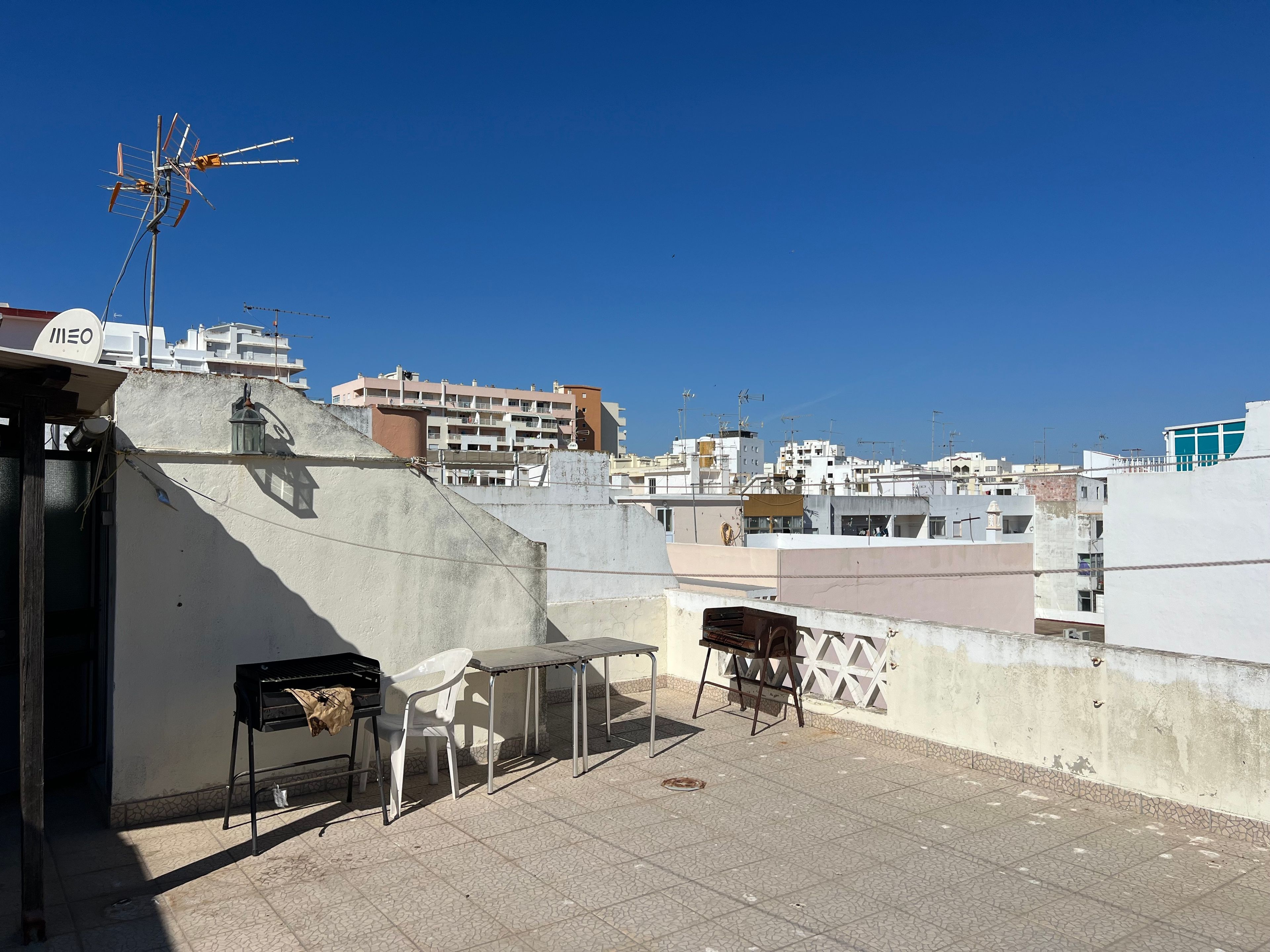 Terraço no telhado com mesa e cadeiras, churrasqueira e vista para edifícios ao redor.