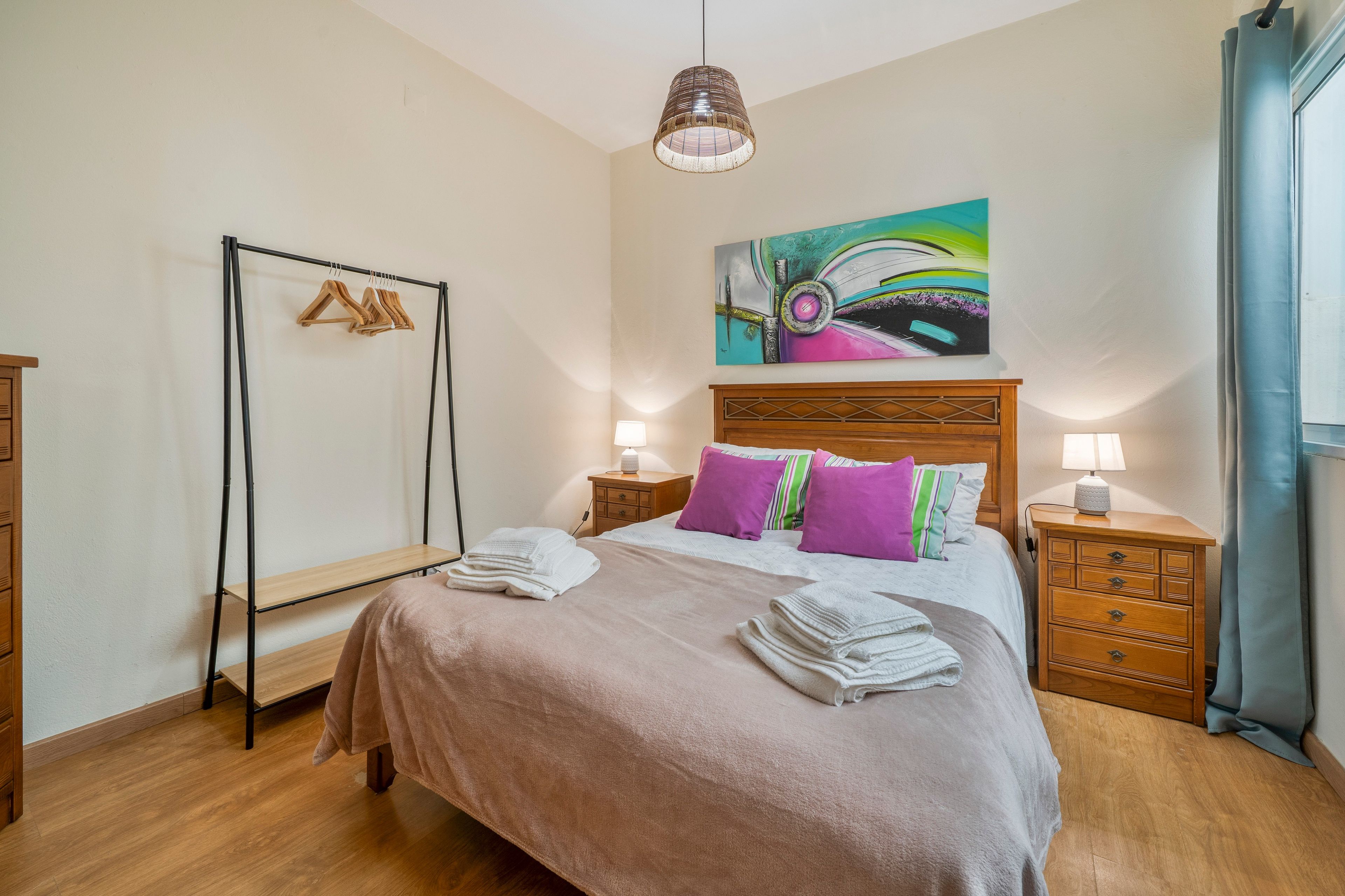 A bedroom with a bed, a bedside table, a hanging light, a painting, and a wardrobe.