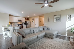 A modern living room with a sectional sofa, a kitchen in the background, and a ceiling fan.