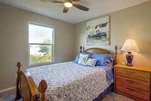 A bedroom with a bed, a nightstand, a lamp, a ceiling fan, a window, and a painting on the wall.
