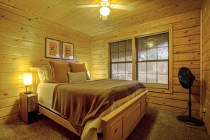 Een slaapkamer in een houten chalet met een bed, nachtkastje, lamp, plafondventilator en jaloezieën.