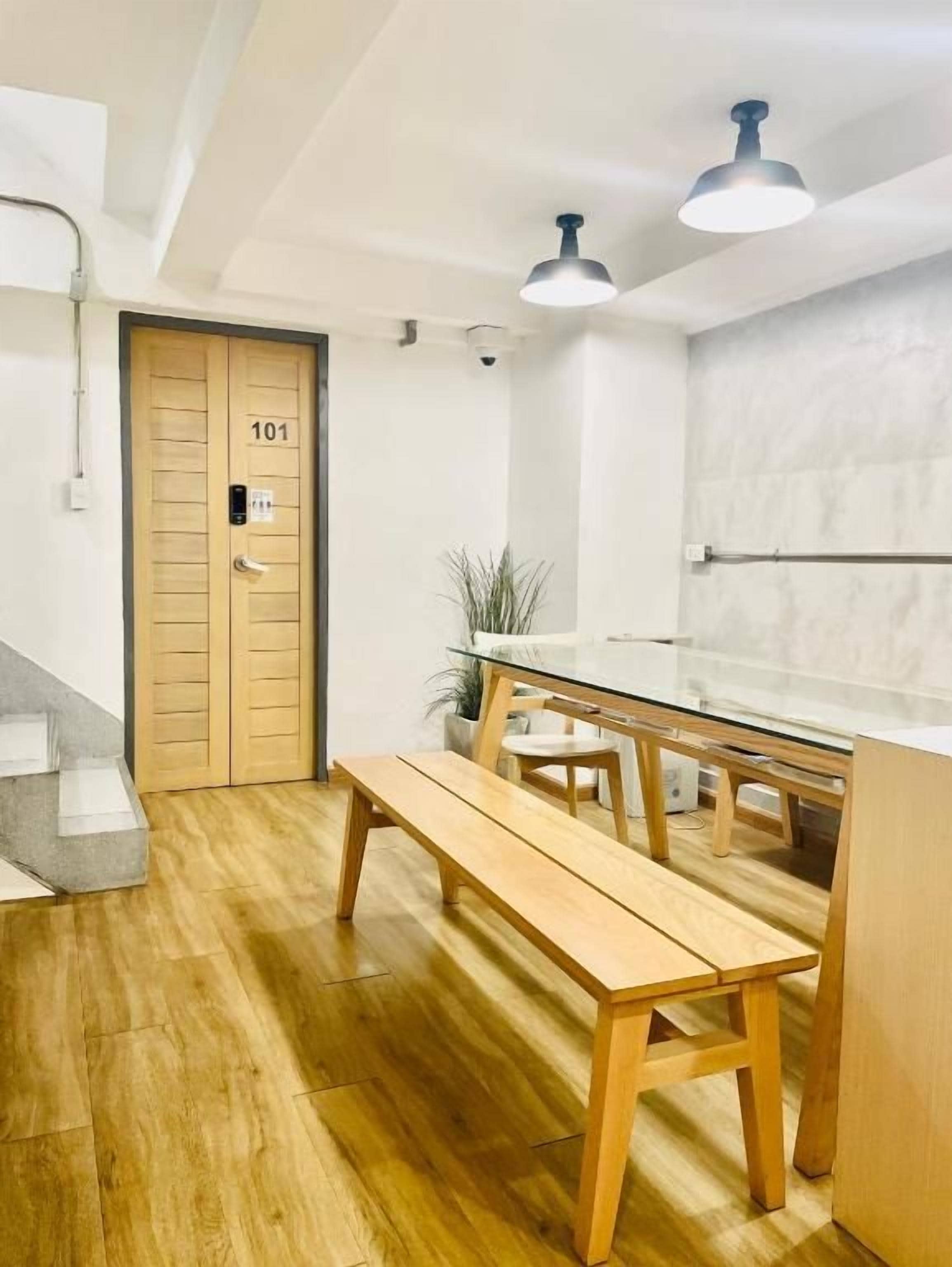 Habitación moderna y minimalista con una puerta de madera identificada con el número '101', una mesa de vidrio y bancos, y una lámpara fijada al techo.