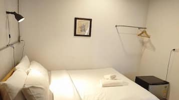 Een enkel bed met witte lakens, een klein nachtkastje, een lamp, een lijst met een afbeelding aan de muur, en een kapstok.