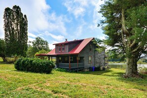 A rustic cabin with a red roof, surrounded by lush greenery and a lake. - On 62-Acre Farm! Waterfront Home w/ Decks & Views (Meadows of Dan)