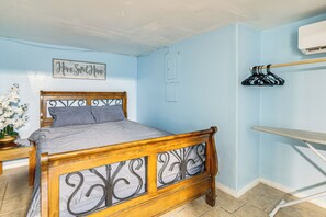 A bedroom with a wooden bed, a nightstand with a plant, a wall-mounted air conditioner, and a framed sign.