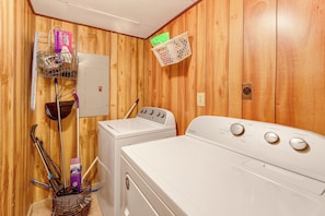 A laundry room with a front-loading washer and dryer, a utility wall with a power outlet, and a basket with cleaning supplies. - Gaston Lake House: Dock, Deck, Game Room! (Bracey)