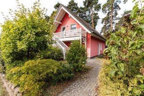 Ett rött hus med en vit balkong, omgivet av grönska och en grusgång.
