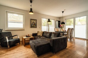 A modern living room with a sectional sofa, a dining area with a table and chairs, and large windows providing natural light. - A gorgeous vacation apartment with a sauna and fireplace near the beach in Röbel Müritz (Röbel/ Müritz)