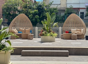 A rooftop terrace with two wicker seating areas, a central planter with plants, and a view of the surrounding buildings. - Qoople Elize A17 - Cozy 1-bedroom apartment in Alanya (Alanya)