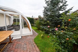 Ein modernes Haus mit einer gewölbten Glasfassade, einer Terrasse mit Tisch und Stühlen sowie einem Garten mit verschiedenen Pflanzen und Blumen.