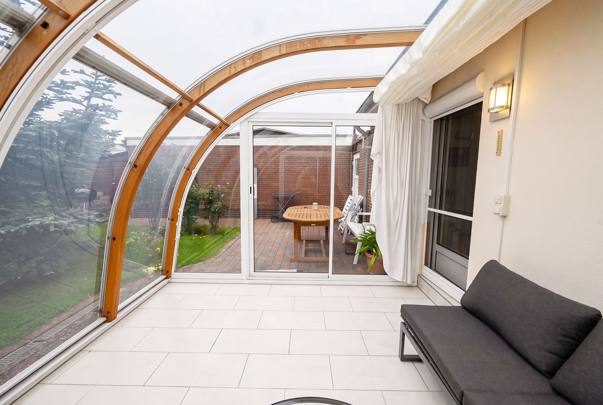 A conservatory with a glass roof, a patio area with a table and chairs, and a grey sofa.