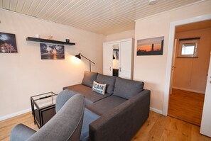 Un salon moderne avec un canapé gris, une table basse en verre et une lampe. Des étagères murales sont fixées au mur et accueillent des objets décoratifs et des tableaux encadrés.