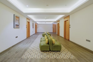 Un pasillo de hotel con alfombra estampada, un sofá verde con cojines, puertas de madera y una lámpara en el techo.