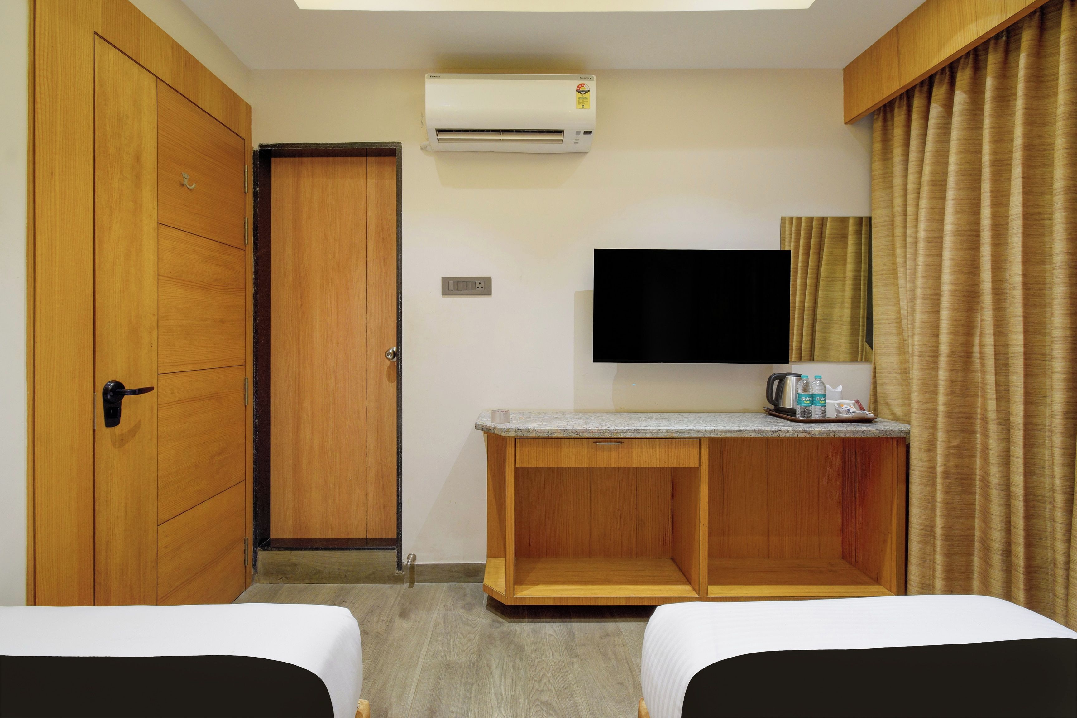 A hotel room with a wooden wardrobe, a desk with a flat-screen TV, two beds, and a ceiling-mounted air conditioner.