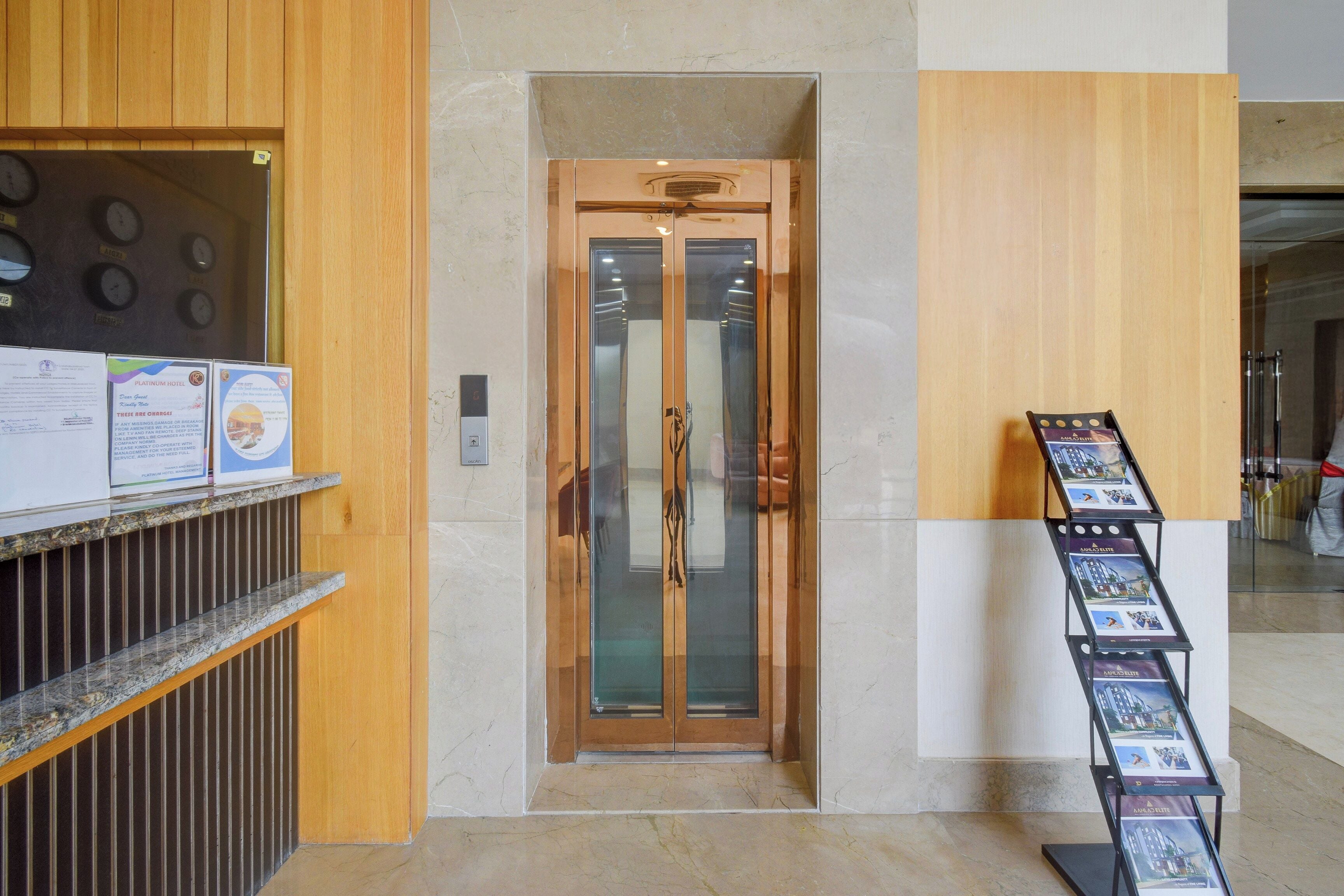 Zona de recepción de un hotel con un mostrador de madera, puerta de ascensor de vidrio y un panel con imágenes de la propiedad.
