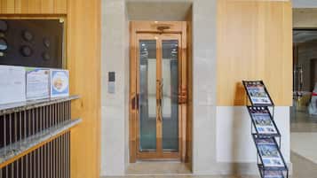 Zona de recepción de un hotel con un mostrador de madera, puerta de ascensor de vidrio y un panel con imágenes de la propiedad.