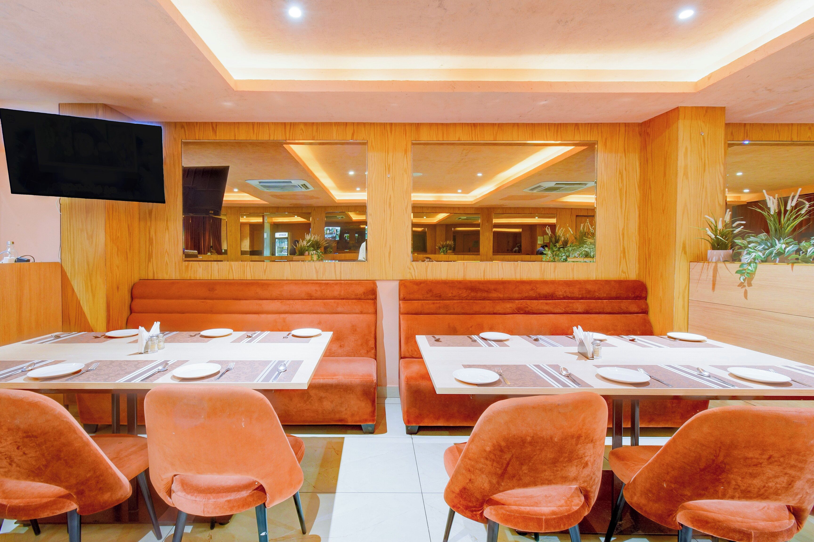 A restaurant with booths and tables, a flat-screen TV mounted on the wall, and potted plants on the side.