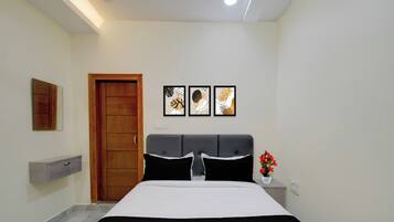 Um quarto com cama, porta de madeira, mesa de cabeceira com um vaso de flores e três quadros na parede.