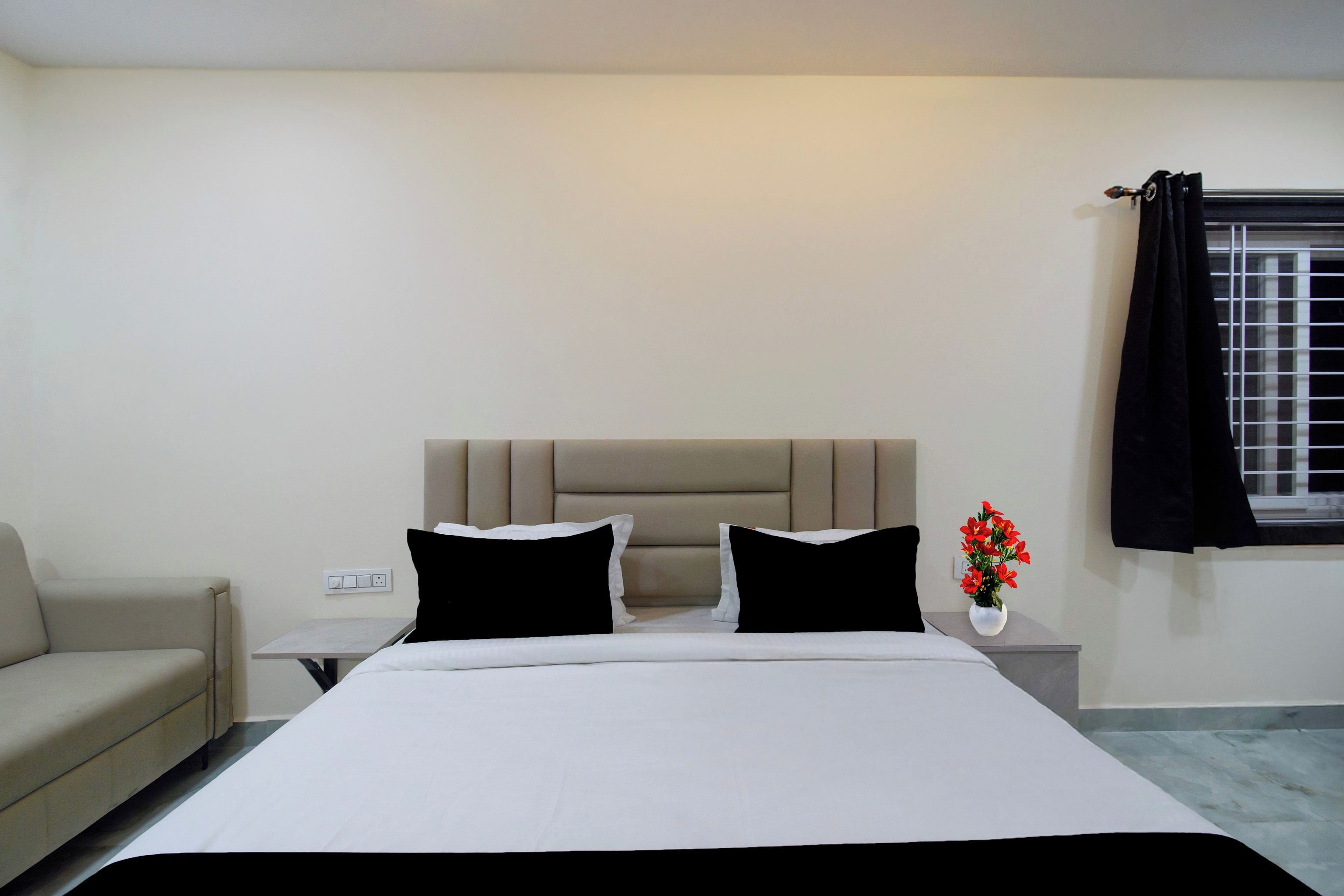 A bedroom with a bed, a nightstand, a lamp, a window with curtains, and a small table with a vase of flowers.
