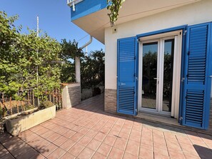 Ein Wohnhaus mit Fliesen-Terrasse, einer blauen Tür und einem Garten mit Zaun und Pflanzen.
