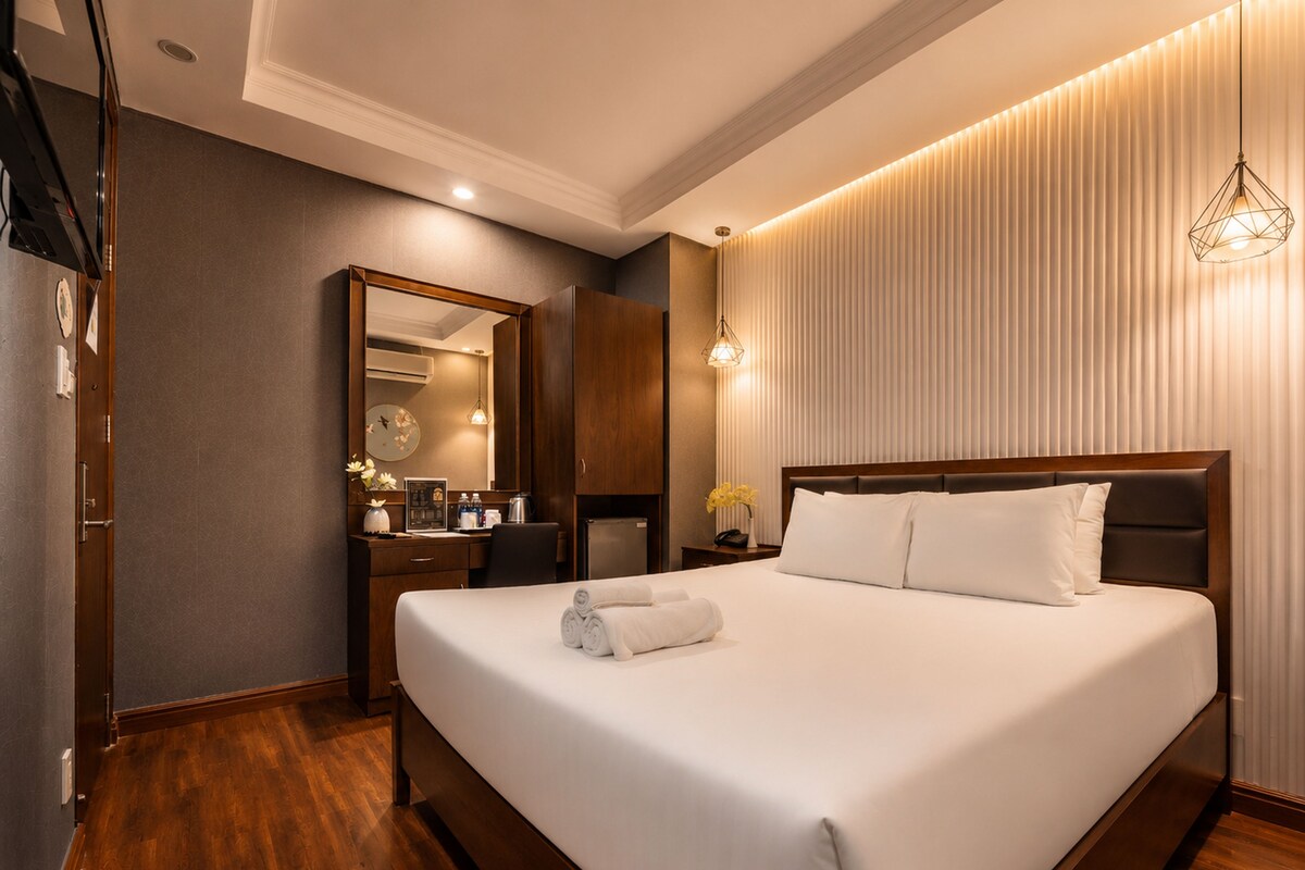 A hotel room with a large bed, a wooden headboard, a mirror, a desk with a chair, and a wall-mounted light fixture.