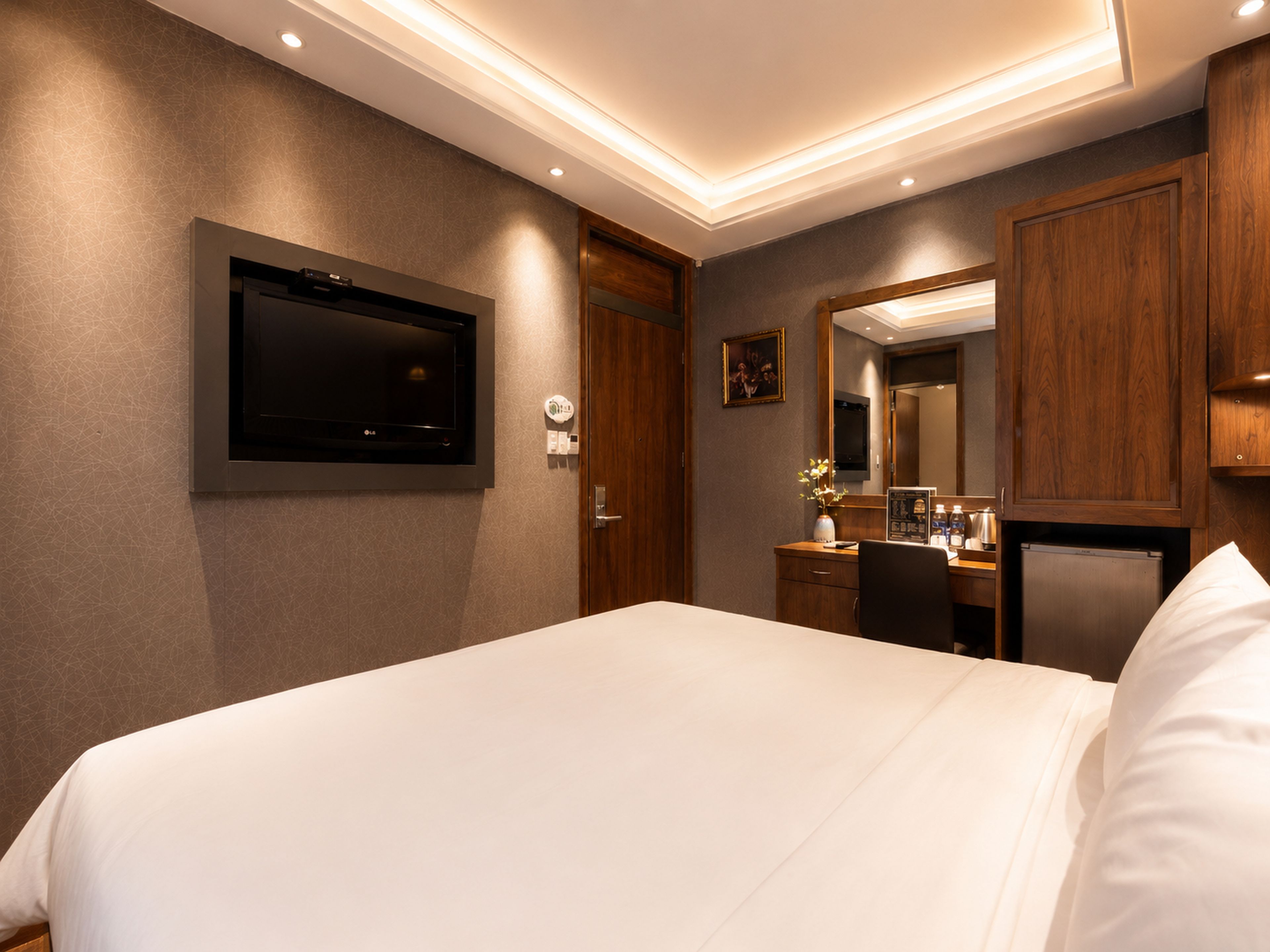 A hotel room with a large bed, a flat-screen TV mounted on the wall, a wooden wardrobe, a desk with a chair, and a mirror above the mirror.