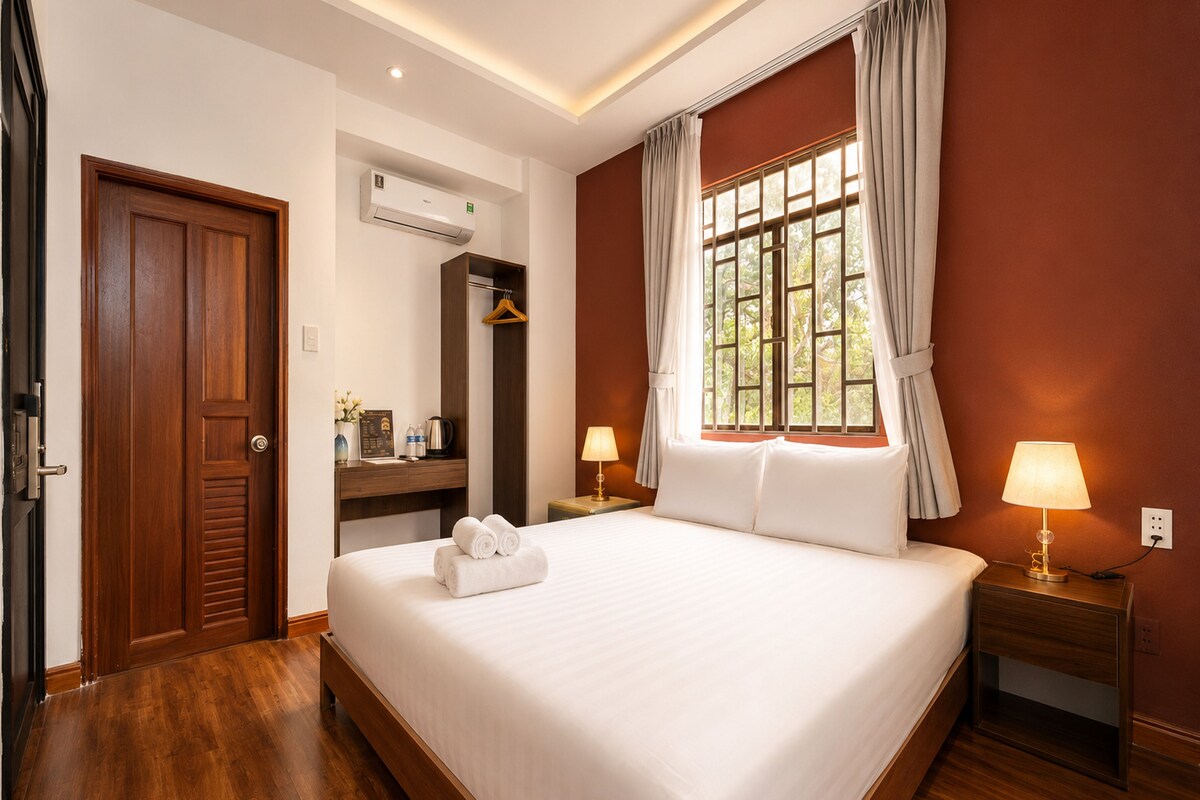 A hotel room with a large bed, a wooden wardrobe, a desk with a chair, a window with curtains, and a bedside lamp.
