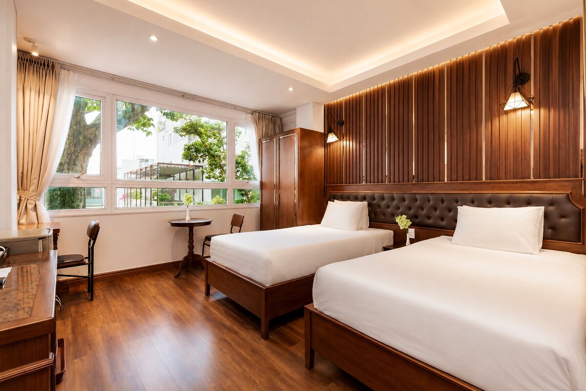 A hotel room with two beds, a desk, a chair, and a window with a view of greenery.