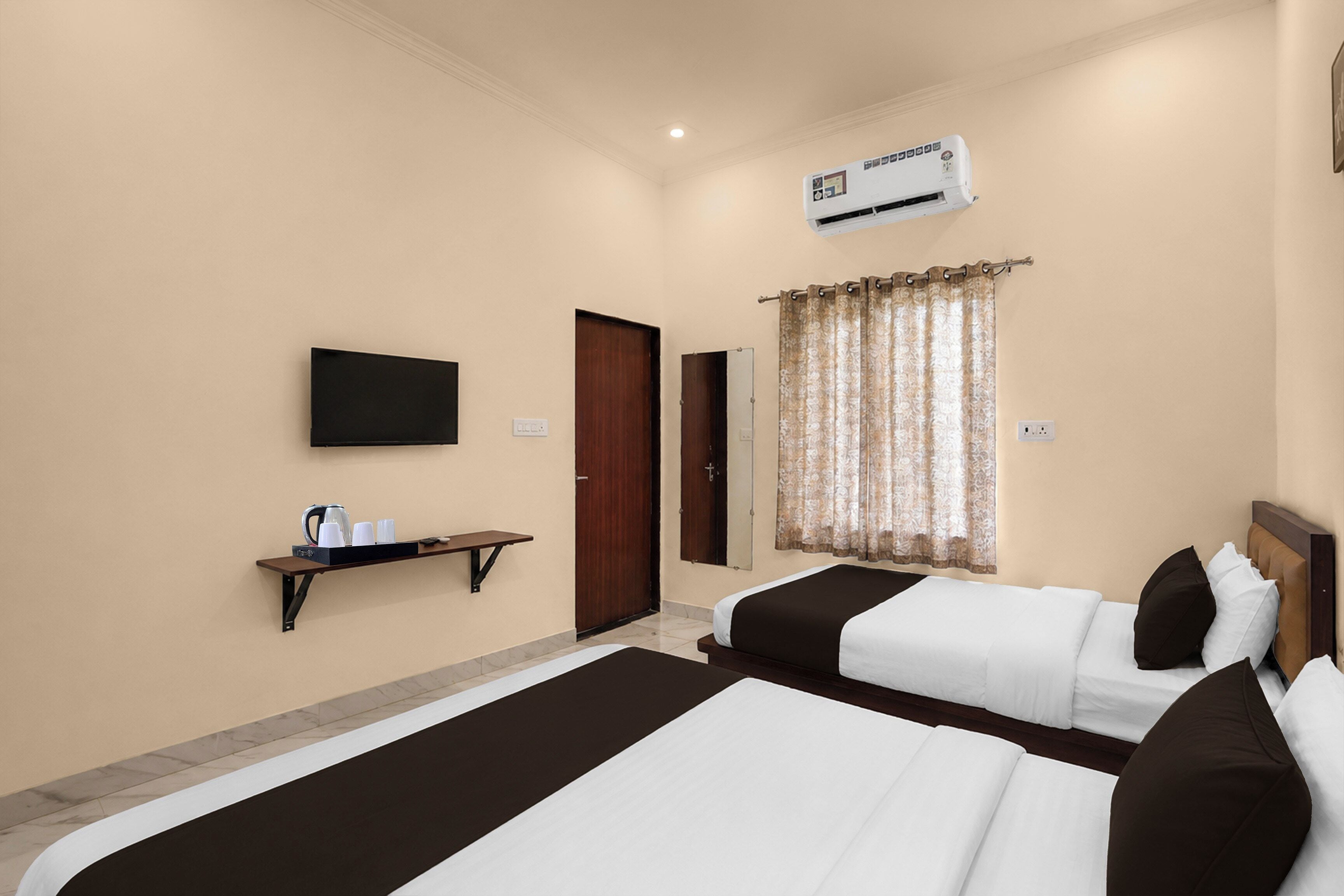 A hotel room with two beds, a flat-screen TV, an air conditioning unit, a small shelf with cups and a kettle, and a window with curtains.