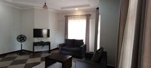 A living room with a flat-screen TV, a sofa, a coffee table, and a standing fan.