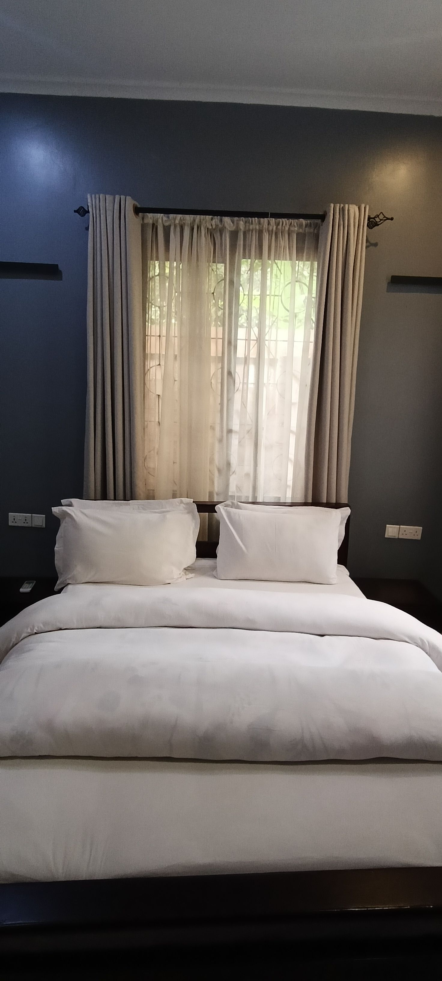Un lit bien fait, avec du linge de lit blanc, dans une pièce aux murs sombres et dotée d’une fenêtre ornée de rideaux légers.