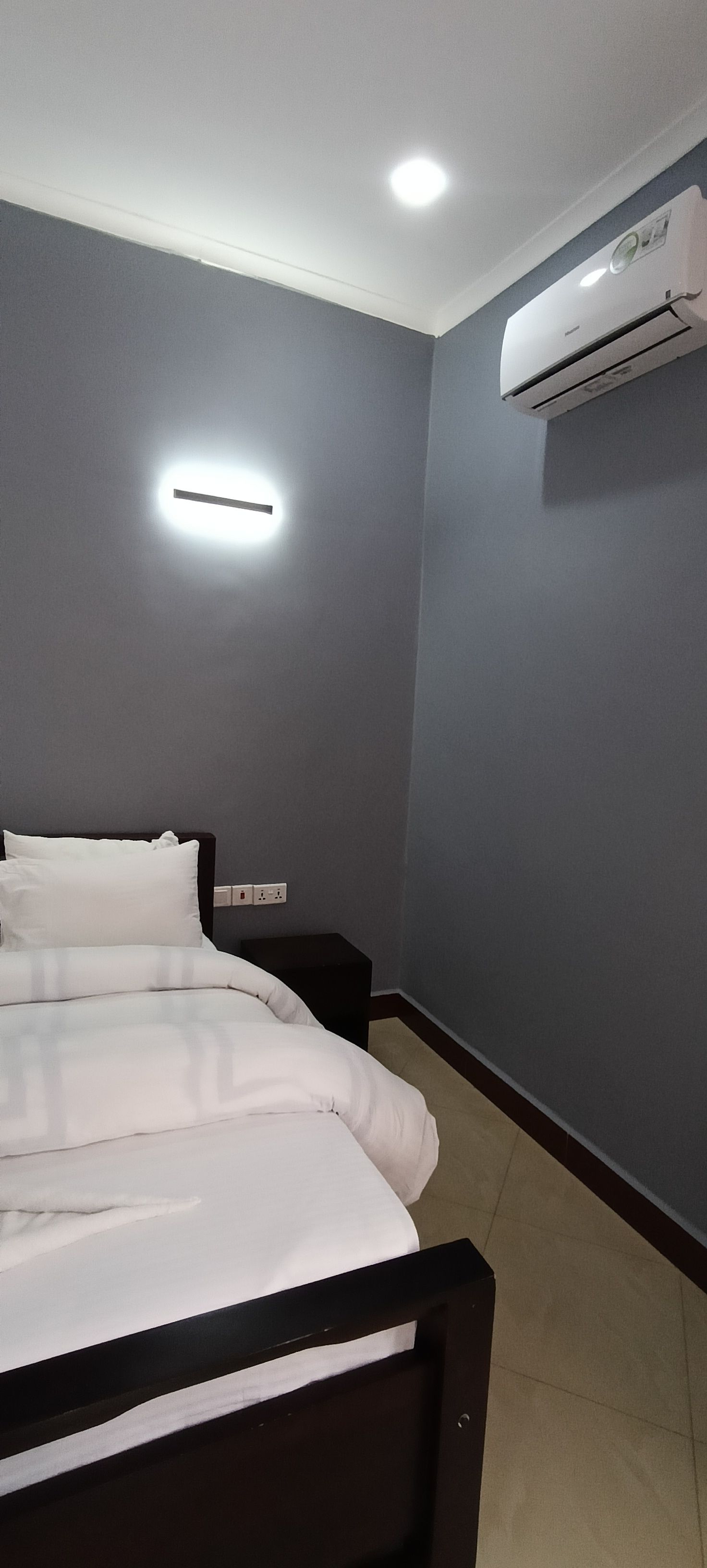 Une chambre à coucher comprenant un lit, une table de chevet, un appareil de climatisation et un luminaire fixé au mur.