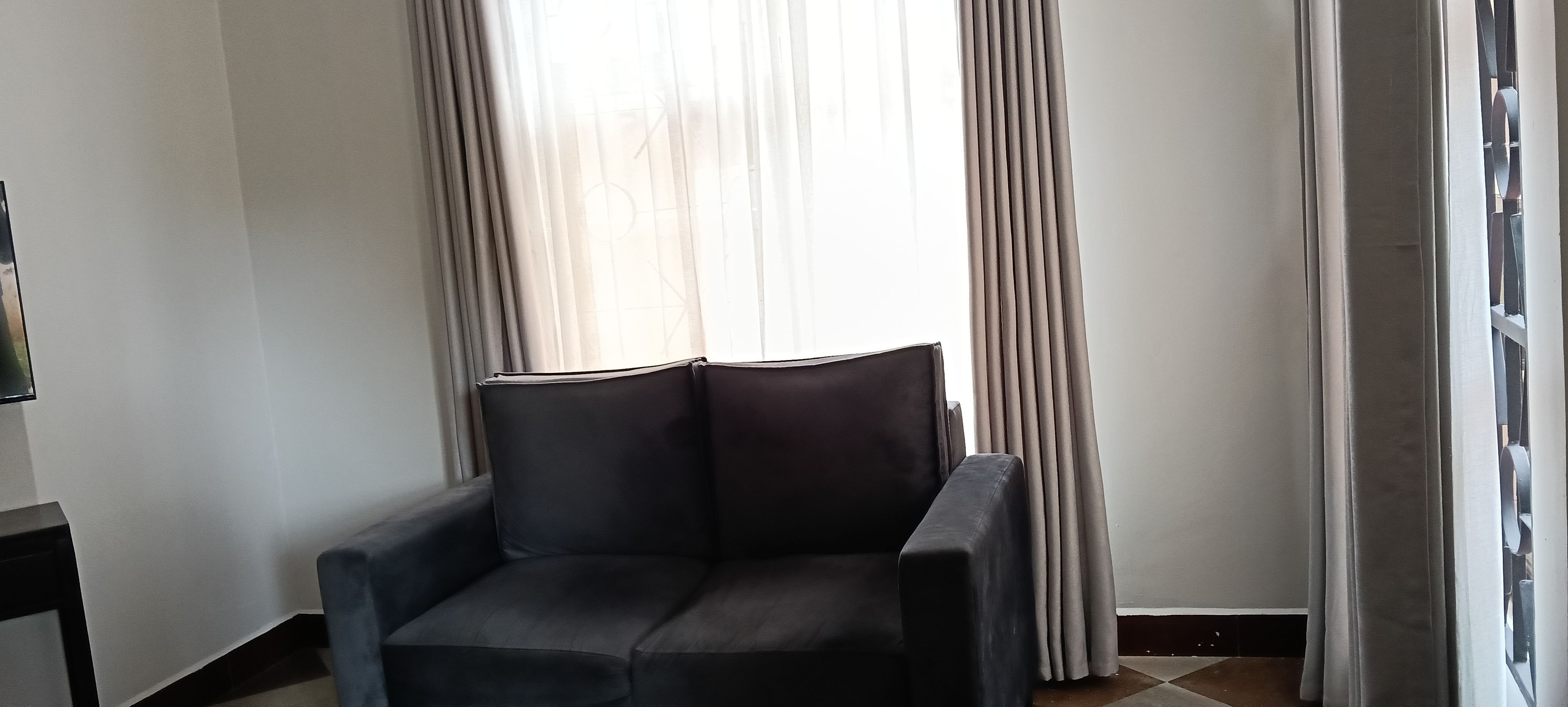 Une pièce d’angle avec un fauteuil de couleur sombre, une fenêtre ornée de rideaux légers et un sol carrelé.