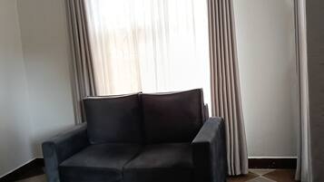 Une pièce d’angle avec un fauteuil de couleur sombre, une fenêtre ornée de rideaux légers et un sol carrelé.