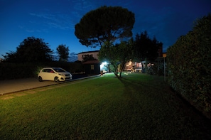 Un barrio residencial con un coche blanco estacionado en la calle, un árbol grande y un edificio con luces encendidas al fondo.