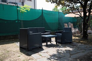 Mueble de mimbre al aire libre sobre baldosas de piedra, valla de malla verde y un árbol con hojas amarillas.