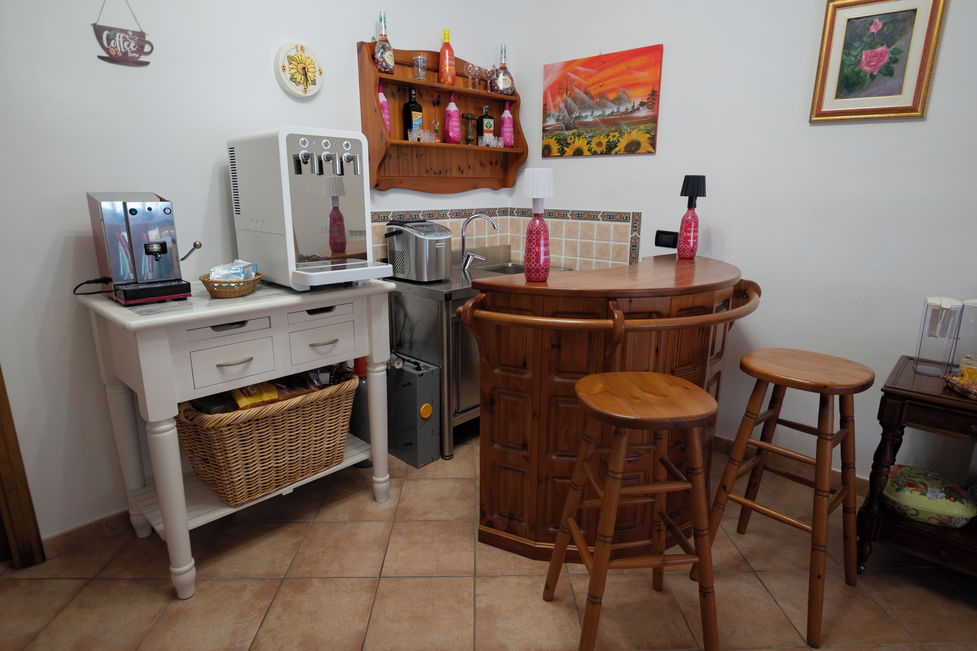 Cocina compacta con cafetera, tostador y lavamanos. Hay dos taburetes y una mesita con una lámpara.