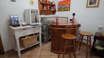Cocina compacta con cafetera, tostador y lavamanos. Hay dos taburetes y una mesita con una lámpara.