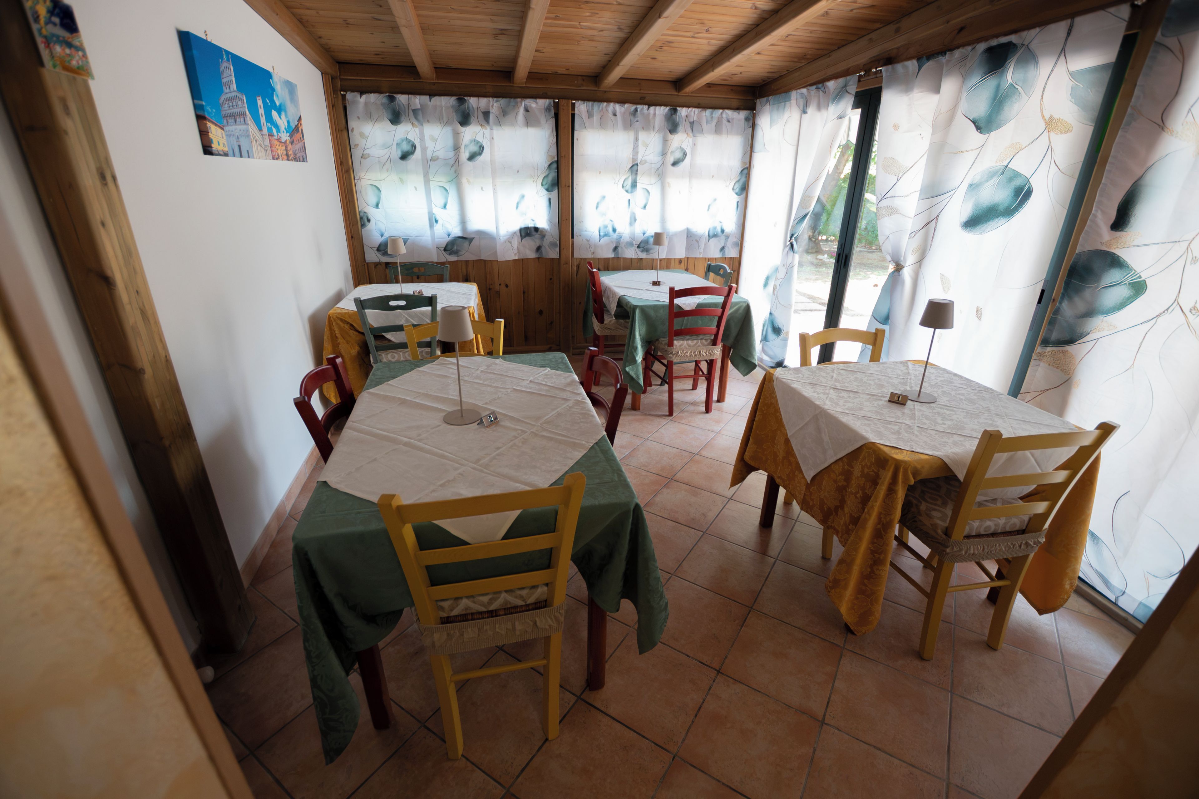 Zona pranzo con tavoli e sedie, travi in legno al soffitto e una finestra con tenda a motivi fogliacei.
