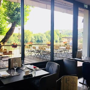 An indoor dining area with a view of a river and greenery. There are tables and chairs arranged for dining, and a menu is visible on the table. - Citotel Chartreuse (Cahors)