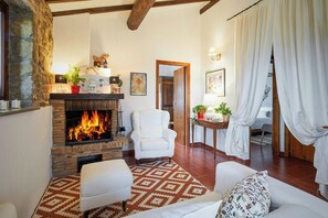 A cozy living room with a fireplace, a white armchair, a patterned rug, and a bed with white curtains. - Cottage surrounded by a romantic garden (Gubbio)