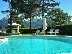 Piscina com espreguiçadeiras e um gazebo, cercada por árvores e montanhas.