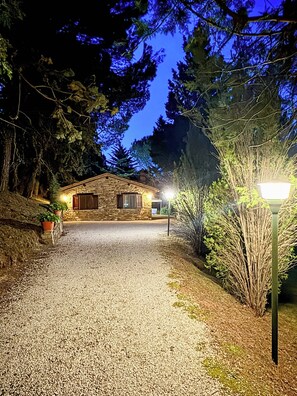A stone house with a gravel driveway, surrounded by trees and a street lamp. - Cottage surrounded by a romantic garden (Gubbio)