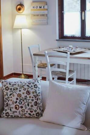 A room with a white sofa, a patterned pillow, a lamp, a table with chairs, and a window with blinds. - Cottage surrounded by a romantic garden (Gubbio)