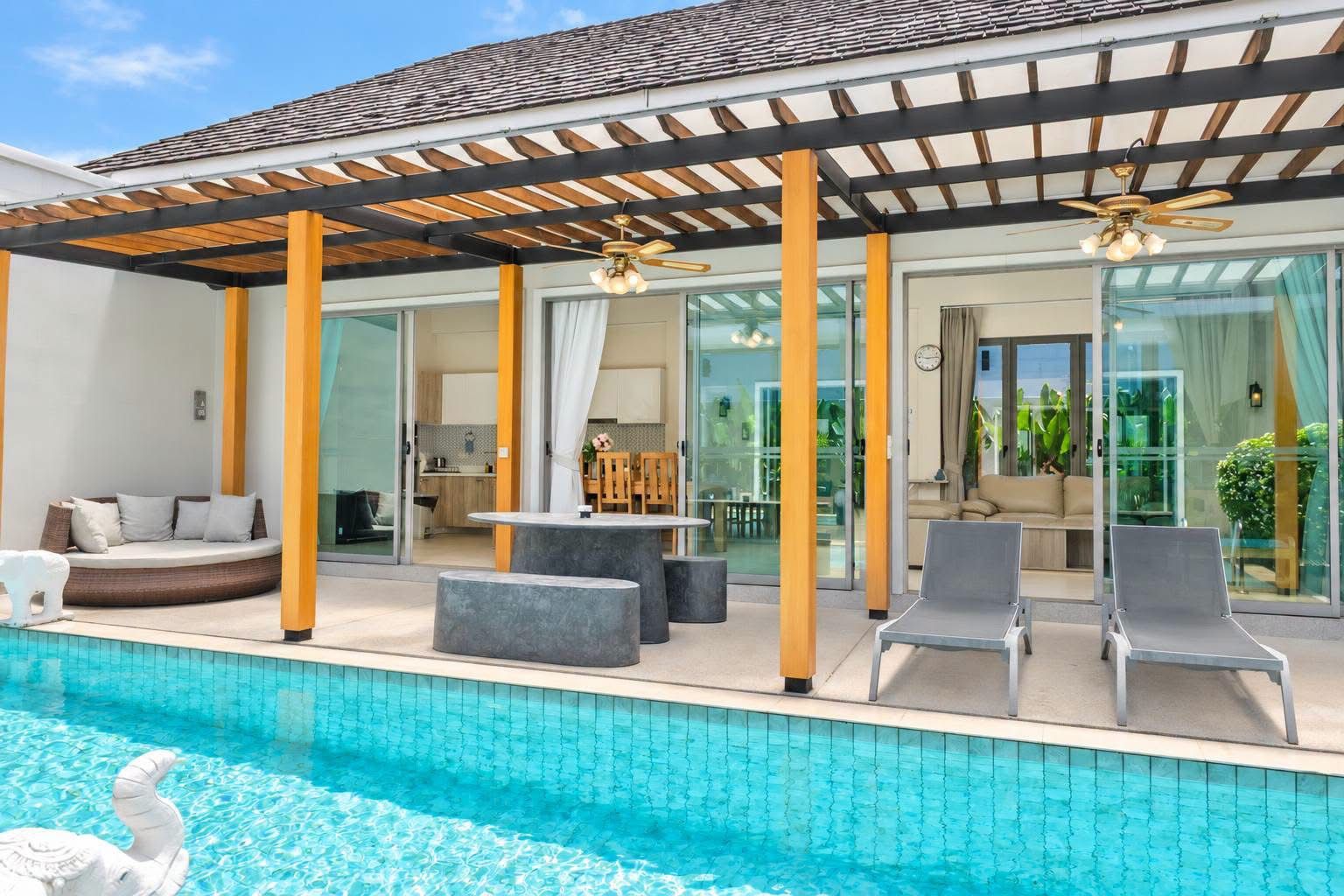 Un área junto a la piscina con zona de estar cubierta, mesa de comedor y sillones reclinables.